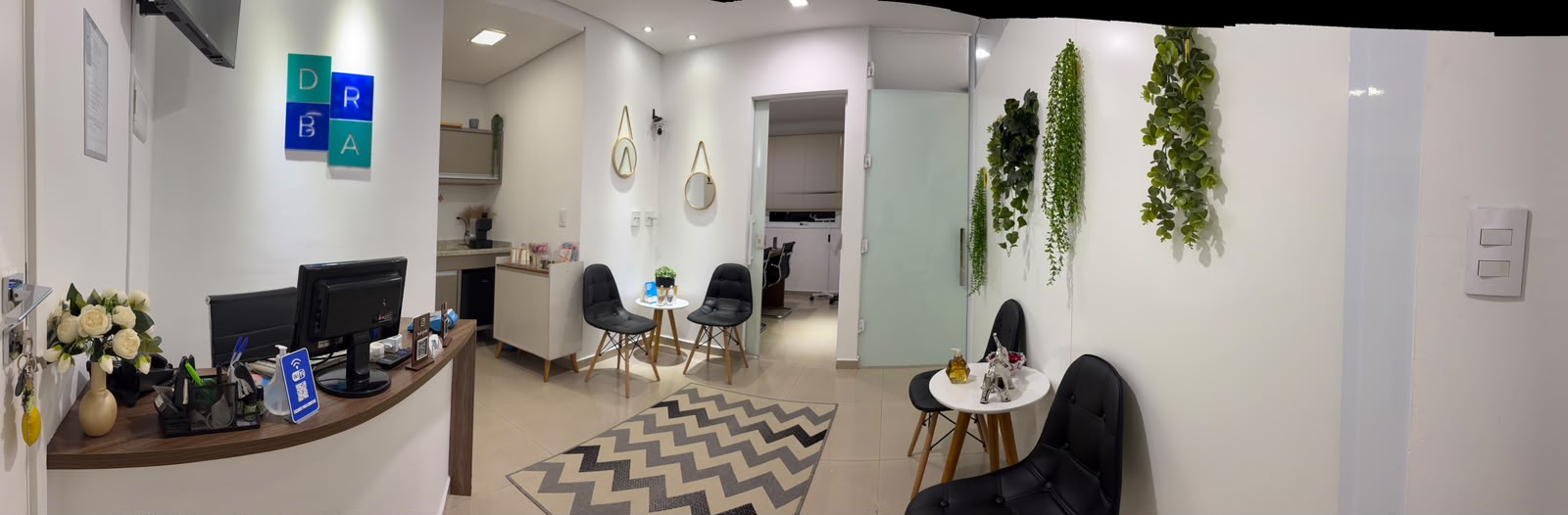 Recepção e sala de espera da Clínica DRBA, com balcão de atendimento, logotipo na parede, cadeiras e plantas